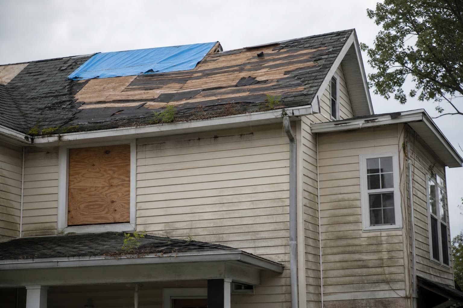 House with a tarp over missing roof shingles and a boarded window