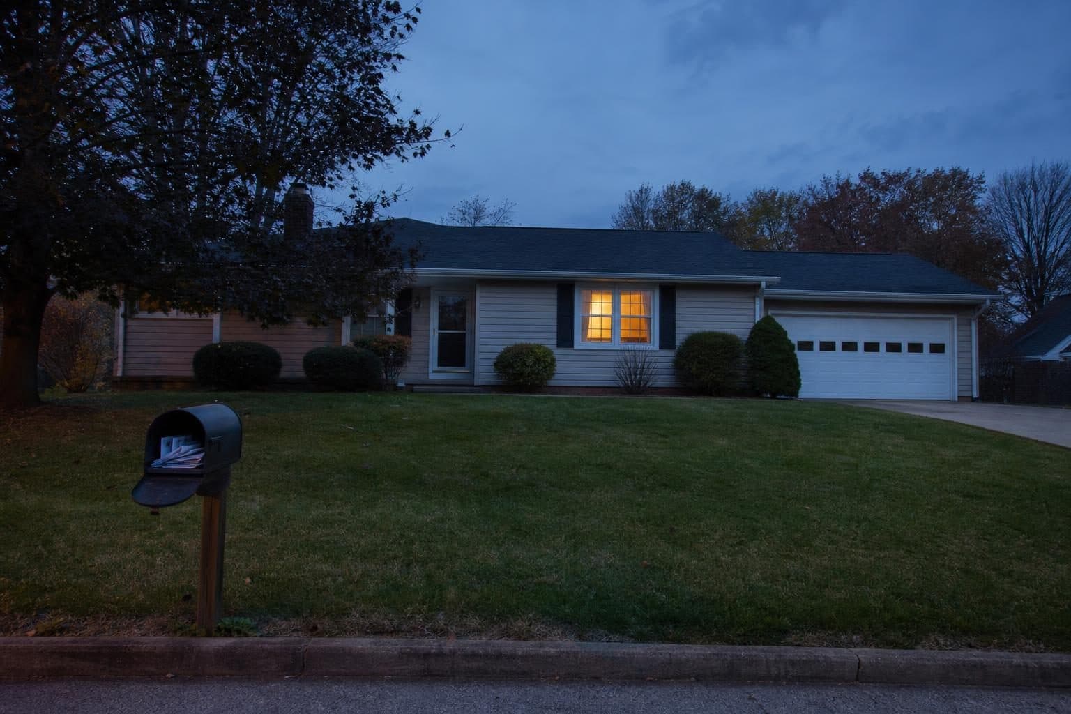 Small ranch house at dusk with a light on and a full mailbox
